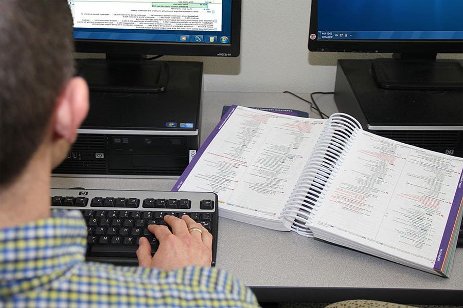 一个人坐在电脑前, 在键盘上打字, with a book of medical coding information open on the desk next to them
